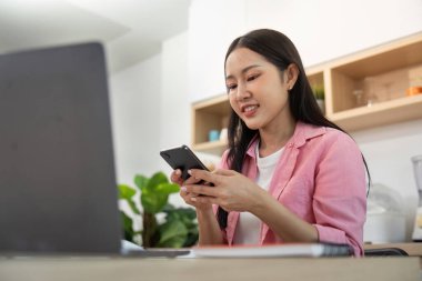 Genç bir kadın akıllı telefonu ve laptopunu kullanarak gülümsüyor, uzaktan kumandalı ev ortamının özünü somutlaştırıyor..