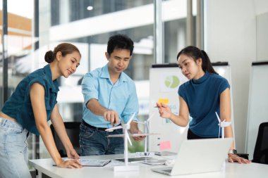 Modern bir ofiste rüzgar türbini modelleri ve dijital araçlar kullanarak sürdürülebilirlik stratejilerini tartışan çeşitli bir ekip.