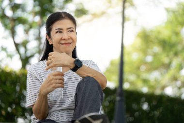 Son sınıf öğrencisi bir kadın parkta bir anlık dinlenmenin keyfini çıkarıyor. Elinde su şişesi tutarken gülümsüyor, sağlığını arttırıyor..