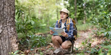 Dişi bir yürüyüşçü canlı bir ormanda çömelir, haritasını dikkatle inceler, doğayla olan bağlantısını ve merakını temsil eder..