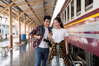 Bir çift telefonlarına bakar, tren istasyonunda beklerken bir sonraki maceralarını planlarlar..