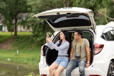 Genç bir çift yolculuk sırasında neşeli anları yakalar, doğada park halindeki arabalarıyla gülümser ve selfie çekerler..