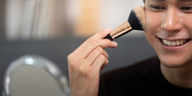 Genç bir adam yanaklarına bir fırçayla allık sürerken gülümsüyor, makyaj rutininin tadını çıkarıyor..