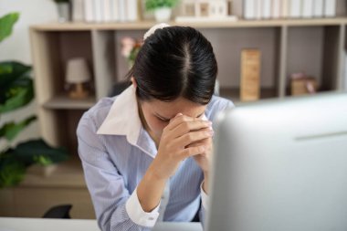 Masasında oturmuş, kurumsal bir ortamda ofis sendromunun etkisini gösteren acı dolu bir ifadeye sahip genç bir kadın kafasını tutuyor..