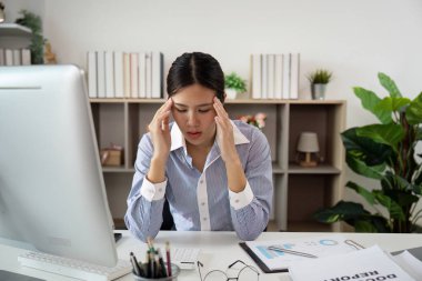 Genç bir kadın şakaklarını tutuyor, masasında çalışırken baş ağrısı çekiyor, ofis sendromunun etkisini vurguluyor..