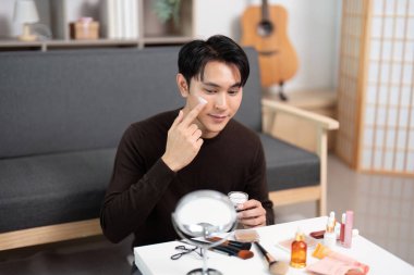 Genç bir adam yüzüne krem sürerek şık bir iç mekanda cilt bakımına konsantre olur..