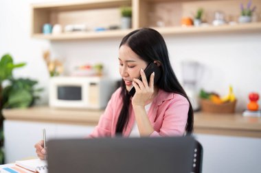 Masasına notlar yazarken bir telefon görüşmesi yapan genç bir kadın. Uzak çalışmada iletişimin vurgulanması.