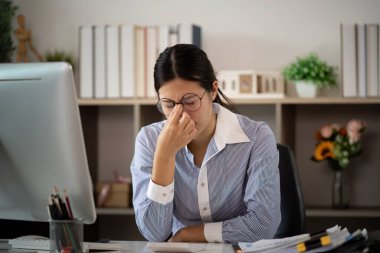 Mavi çizgili gömlekli bir kadın modern bir ofiste bilgisayarında çalışırken stres ve yorgunluk belirtileri gösteriyor..