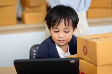 Genç bir çocuk, ev ofisindeki kutularla çevrili bir şekilde tabletine odaklanarak modern aile yaşamında oyun ve verimliliğin karışımını sergiliyor..