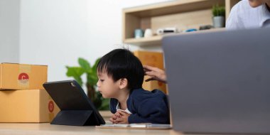 Genç bir çocuk bir tabletle meşgul. Annesi, uzak çalışmalardaki aile dinamiklerini gösteren bir ev ofisinde görevlerini yönetiyor..