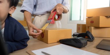 Bir anne, çocuğu izlerken bir nakliye kutusunu mühürlemek için bantla ebeveynlik ve ev işi görevlerinin karışımını vurgular..