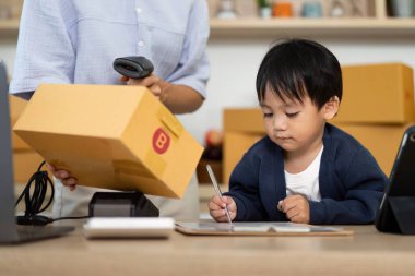 Genç bir çocuk, annesinin bir ev ofisindeki iş ve aile hayatının uyumunu gösteren bir paketi tararken yazmaya odaklanır..