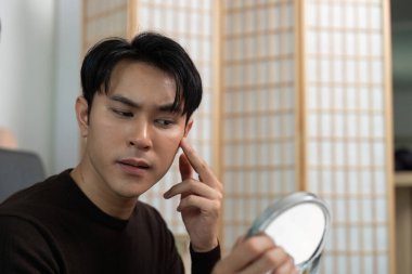 Genç bir adam derisini aynada yakından inceleyerek cilt bakımına odaklanır. Kişisel bakım ve tımarın altını çiziyorum..