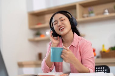 Evde neşeli bir kadın, kulaklıkla müzik dinliyor ve kahvesinin tadını çıkarıyor, rahat bir çalışma ortamını yansıtıyor..