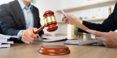 Bir avukat belgeleri incelerken bir tokmak kaldırıyor, modern ofis ortamında otoriteyi ve hukuki süreci vurguluyor.