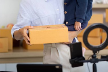 Bir anne çocuğunu taşırken bir paket tutar, aile hayatı ve iş hayatının karışımını bir ev ofisinde gösterir..