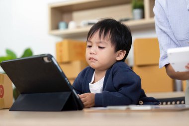 Genç bir çocuk, ev ofisindeki bir tablete odaklanır. Teknoloji ve aile hayatının karışımını vurgular..