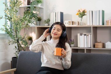 Genç bir kadın kulaklıkla müzik dinliyor ve kahvesini yudumluyor, şık oturma odasında rahat bir anın tadını çıkarıyor..