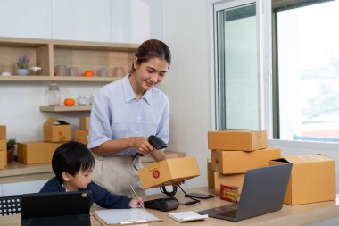 Bir anne, çocuğu bir tabletle uğraşırken paketleri tarar ve aile ve girişimcilik dengesini bir ev ofisinde gösterir..