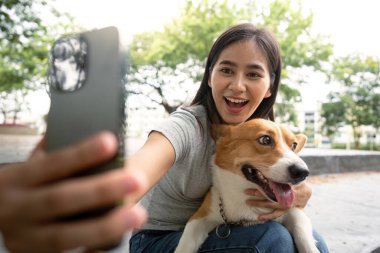 Genç bir kadın, canlı bir park ortamında evcil hayvan sahibi olmanın mutluluğunu kutlayan korgisiyle neşeli bir selfie çekiyor..