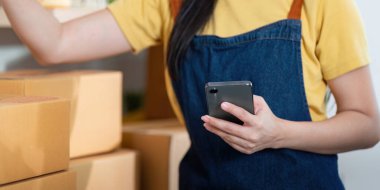 Bir girişimci, etrafı nakliye paketleriyle çevriliyken akıllı bir telefondan envanteri kontrol eder ve modern iş uygulamalarını ön plana çıkarır..