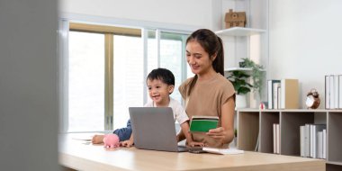 Gülümseyen bir anne oğlunun dijital finans öğrenmesine yardım ederken modern bir ev ofisinde kumbarasını kullanıyor..