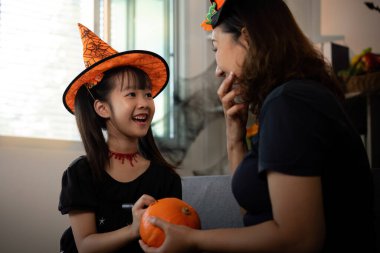 Cadı şapkalı genç bir kız ve annesi yaratıcı bir cadılar bayramı aktivitesinden zevk alıyorlar, gülümseyerek balkabaklarını boyuyorlar ve gülüyorlar..