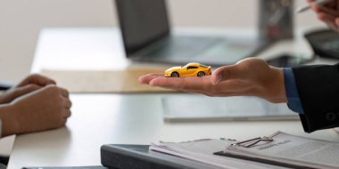 Bir sigorta acentesi, bir müşteriye danışma sırasında sigorta seçeneklerini açıklarken oyuncak bir arabayı elinde tutar..