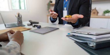 Bir sigorta acentesi elinde oyuncak bir araba tutuyor ve bir müşteriye sigorta poliçesinin detaylarını anlatıyor..