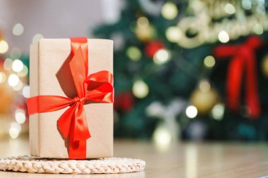 A neatly wrapped gift box with a red ribbon, set against a blurred Christmas tree filled with lights and ornaments.