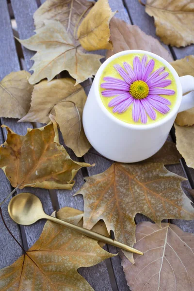 Golden milk in a white cup decorated with a pink daisy. Anti-inflammatory drink. Golden milk with autumn leaves background.