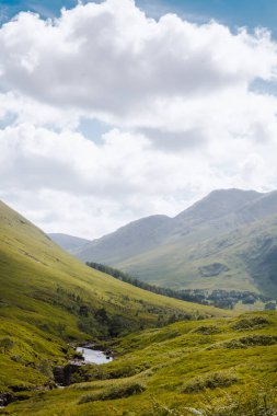 Glencoe Vadisi 'nin geniş manzarası kıvrımlı bir nehir, yeşil dağlar ve kabarık bulutlarla dolu parlak mavi bir gökyüzü..