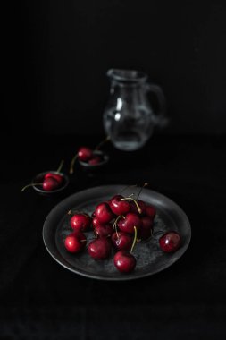 Arka planda cam sürahi olan bir tabak taze kiraz, siyah arka planda klasik bir natürmort oluşturur..