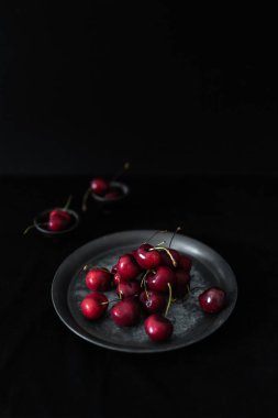 Bir tabak taze, olgun kiraz, siyah arkaplanlı koyu dokulu bir tabakta güzelce sıralanmış.
