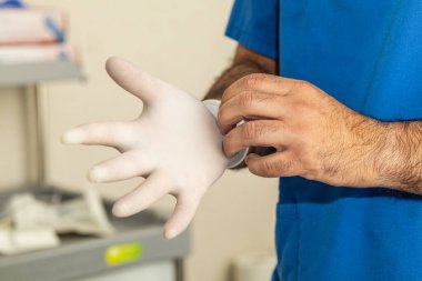 Mavi önlüklü bir sağlık uzmanı cerrahi eldiven takıyor, tıbbi bir ortamda el hijyeni ve güvenliği vurguluyor..