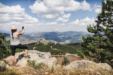 Şapkalı ve çizgili atletli bir kadın akıllı telefonuyla bir dağın tepesinde fotoğraf çeker ve Lagunas de Neila 'nın nefes kesici manzarasını yakalar.