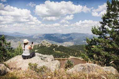 Şapkalı ve çizgili atletli bir kadın akıllı telefonuyla bir dağın tepesinde fotoğraf çeker ve Lagunas de Neila 'nın nefes kesici manzarasını yakalar.