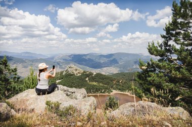 Bir kadın kayanın üzerinde oturuyor, akıllı telefonuyla selfie çekiyor ve Lagunas de Neila 'nın nefes kesici manzarasının tadını çıkarıyor..