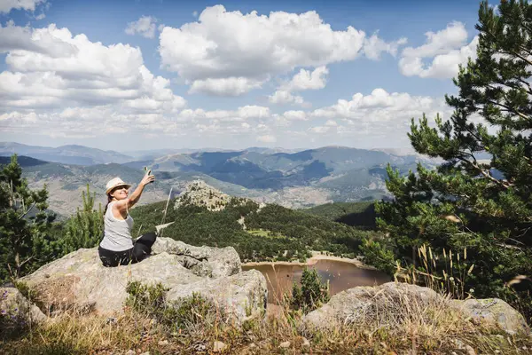 Çizgili askılı tişörtlü ve şapkalı bir kadın bir dağın tepesinde selfie çeker ve Lagunas de Neila 'nın büyüleyici manzarasını yakalar..