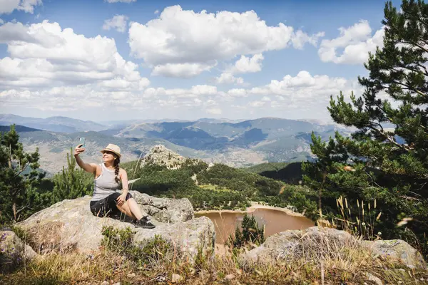 Çizgili askılı tişörtlü ve şapkalı bir kadın bir dağın tepesinde selfie çeker ve Lagunas de Neila 'nın büyüleyici manzarasını yakalar..