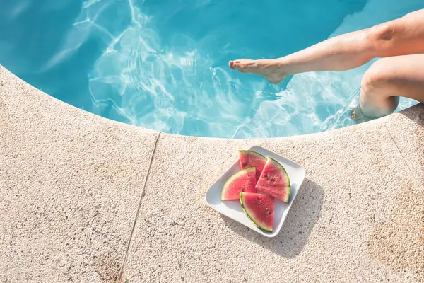 stock image A leg dipped in the pool while lounging next to a plate of watermelon slices, highlighting a moment of summer relaxation.