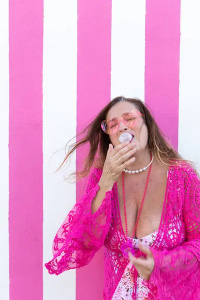 Pembe elbiseli neşeli bir kadın pembe ve beyaz çizgili bir duvara sakız üflüyor, eğlenceli, renkli bir sahne yaratıyor..