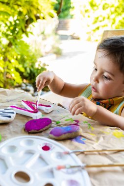 A young boy enjoys painting with watercolors in the garden, a perfect outdoor activity that encourages creativity and healthy development. clipart