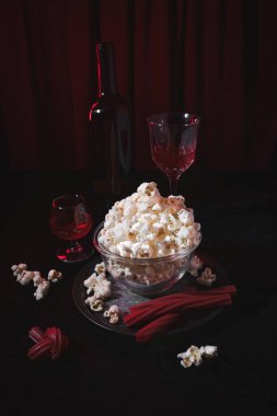 A still life composition with a bowl of popcorn, red drinks in glasses, red licorice sticks, and a dark background. The scene suggests a cozy movie night or snack time with a cinematic feel. clipart