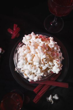 A flat lay view of popcorn in a bowl, red drinks in glasses, and red licorice sticks on a dark background. The arrangement conveys a cozy movie night concept, perfect for a cinematic snack setup. clipart