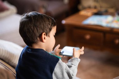 Üç yaşında bir çocuk kanepede oturuyor, elinde bir akıllı telefon tutuyor, çocukların teknolojiyle olan ilişkilerini gösteriyor.