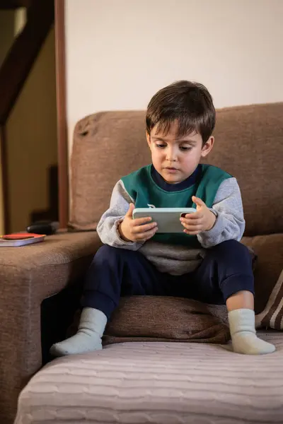 Üç yaşında bir çocuk kanepede oturuyor, elinde bir akıllı telefon tutuyor, çocukların teknolojiyle olan ilişkilerini gösteriyor.