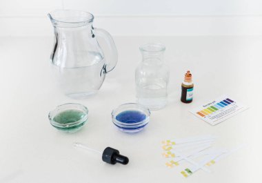 Renk göstergesi düşüşlerini de içeren bir su pH test kitinin fotoğrafları ve evde veya eğitim ortamlarında içme suyunun asitlik veya alkalikliğini analiz etmek için kullanılan bir karşılaştırma grafiği.