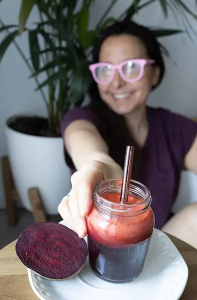 Arka planda bir kadın içmek için uzanırken taze pancar püresi ön planda oturur. Görüntü, sağlıklı yaşam, doğal beslenme ve bitkisel alışkanlıklar yoluyla sağlıklı günlük yaşam kavramını yakalıyor..