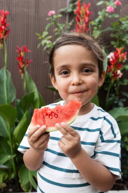 Çizgili gömlek giyen üç yaşındaki neşeli bir çocuk büyük bir dilim kırmızı karpuzun tadını çıkarıyor..